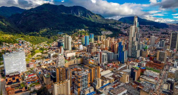 Aerial view of Bogotá’s dense skyline with modern skyscrapers backed by lush Andean hills under patchy clouds.