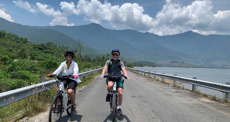 Two cyclists ride along a coastal road with lush mountains and blue sky in central Vietnam.