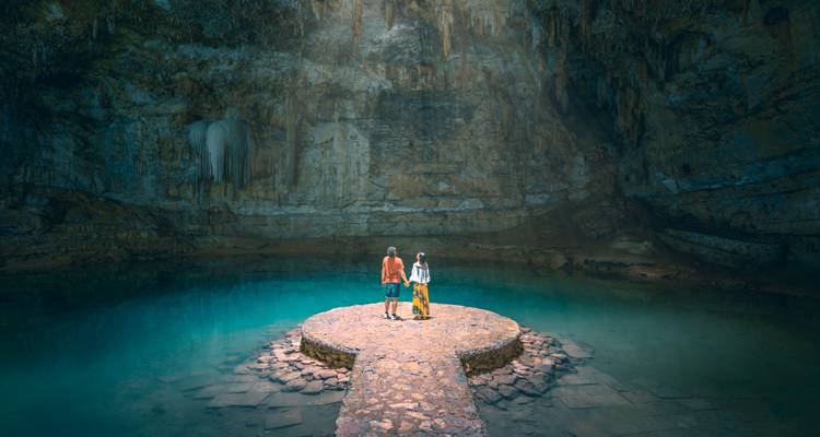 Deux voyageurs se tiennent sur une plateforme de pierre au-dessus d'une eau d'un bleu éclatant à l'intérieur d'une spectaculaire grotte de cénote.