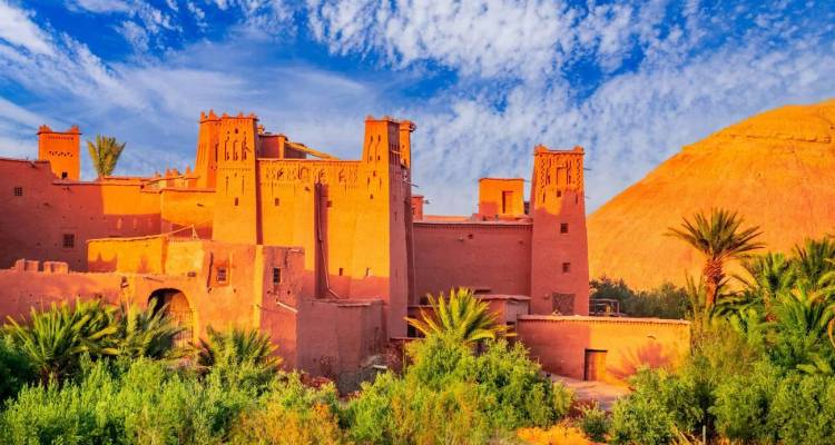 Erdfarben getönte Kasbah von Ait Benhaddou vor einem lebhaften blauen Himmel mit grünen Vordergrundbäumen.