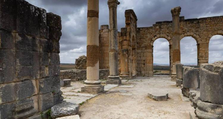 Verwitterte Steinbögen und Säulen der Ruinen von Volubilis unter einem düsteren grauen Himmel.