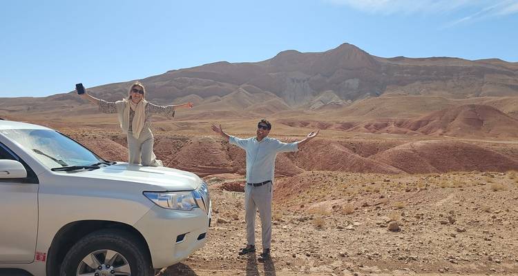 Deux voyageurs célèbrent au-dessus et à côté d'un 4x4 blanc face aux collines désertiques rouges du Maroc.
