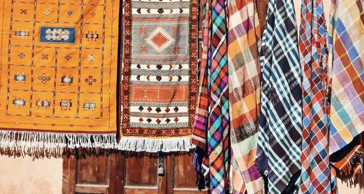Des tapis et des couvertures marocains vibrants sont suspendus en rangées colorées contre une porte en bois.