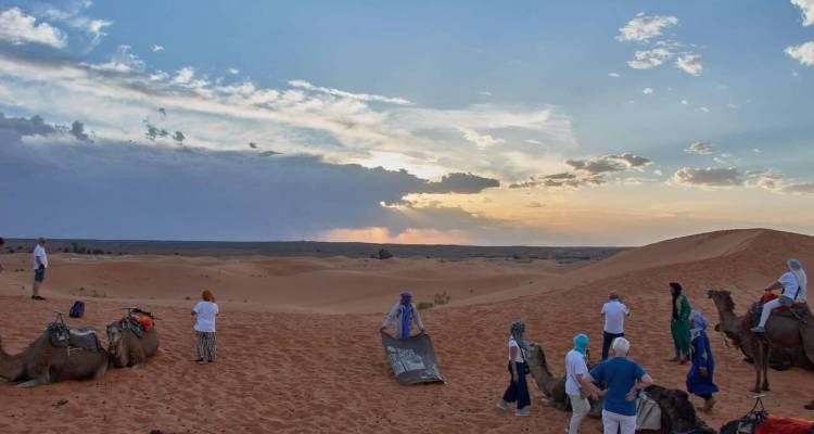 Les voyageurs et les chameaux se rassemblent sur des dunes orange au crépuscule avec des nuages dramatiques à l'horizon.