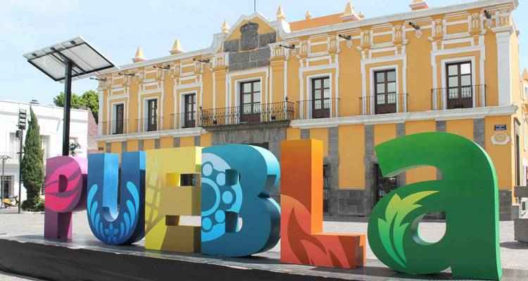 Große bunte Buchstaben, die PUEBLA buchstabieren, stehen vor einem reich verzierten gelben Kolonialgebäude auf einem sonnigen Platz.