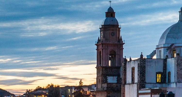 Ein Glockenturm und eine weiße Kuppel zeichnen sich gegen einen dramatischen blaugrauen Abendhimmel in einer mexikanischen Stadt ab.