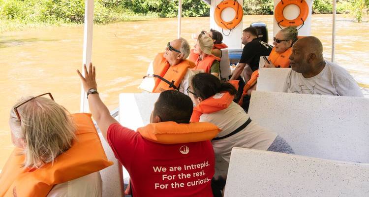 A boatload of visitors wearing orange life vests cruises down a muddy rainforest river.