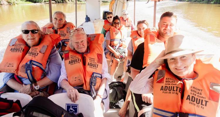 Tourists in life vests smile and wave aboard a covered boat on a jungle waterway.