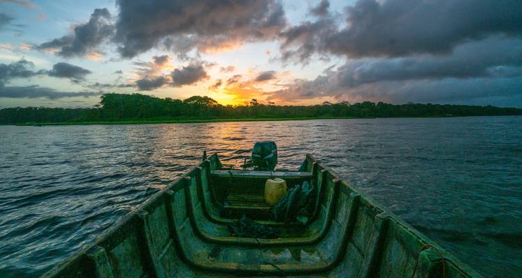 Aussicht vom Bug eines kleinen Holzboots, das bei lebendigem Sonnenuntergang über einen breiten Fluss oder eine Lagune fährt.