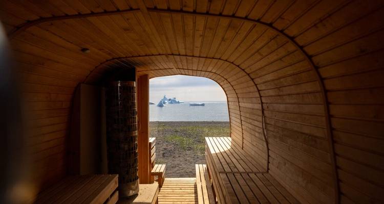View from inside a wooden seaside sauna looking out to icebergs drifting in calm water.