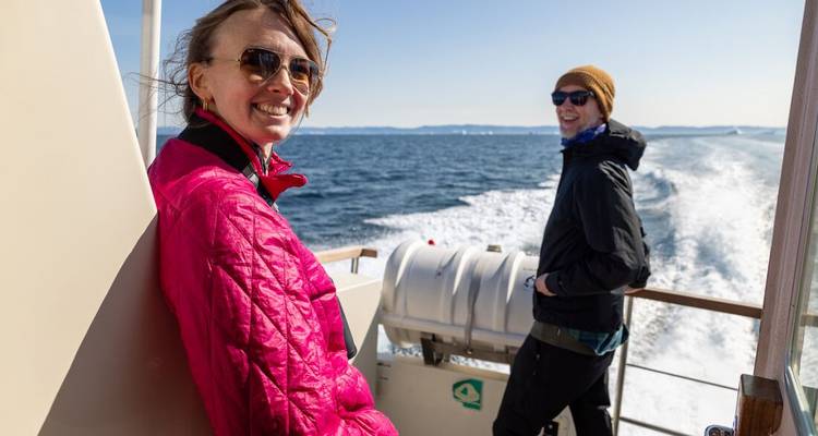 Two travellers smile on the deck of a fast-moving boat leaving a foamy wake.