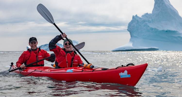 Two paddlers in red drysuits kayak past a towering iceberg on calm Arctic waters.