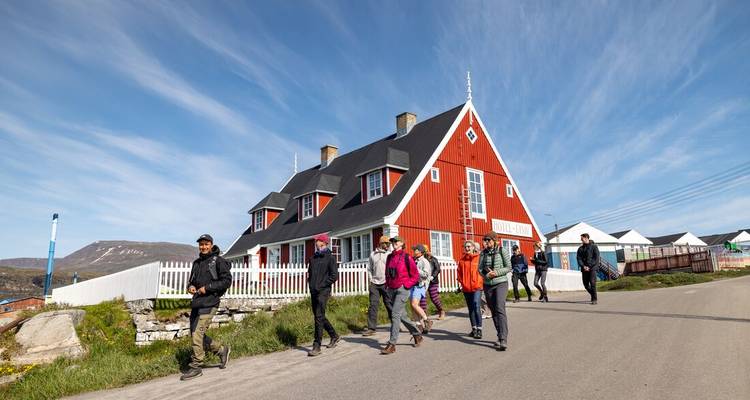 Travellers walk along a road past a traditional red-painted Greenlandic house under clear skies.