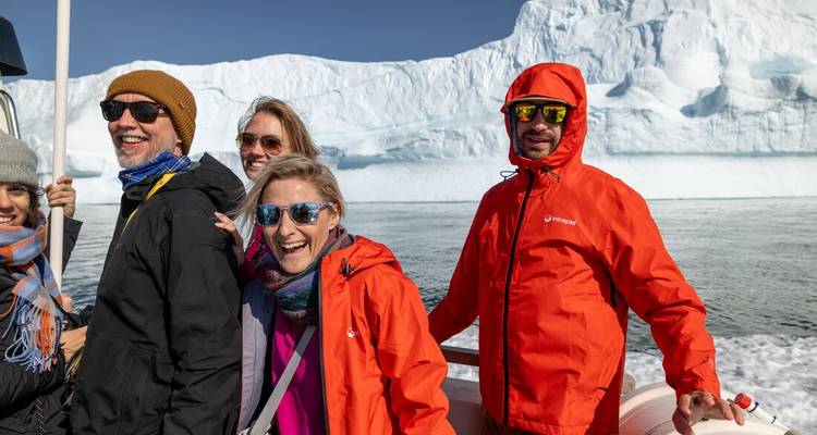 A group on a small boat enjoys sunshine with a massive iceberg backdrop.