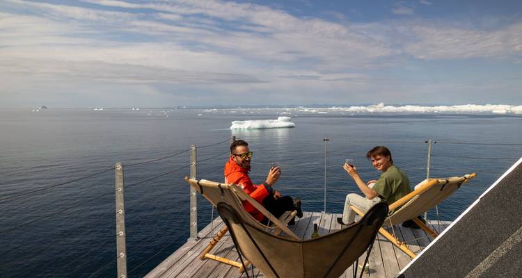 Two guests relax in deck chairs on a wooden terrace overlooking a sea filled with icebergs.