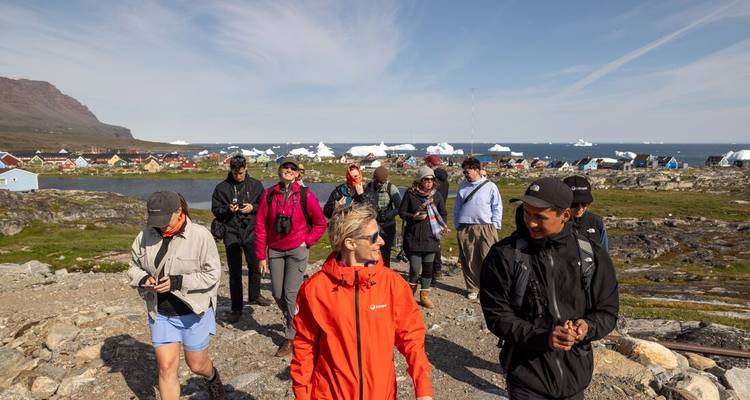 Travellers walk through a coastal Greenland village with icebergs floating offshore.