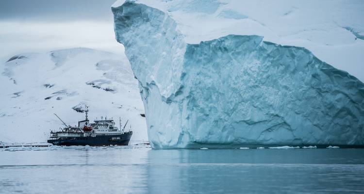 Un immense iceberg bleu se dresse au-dessus des eaux arctiques calmes tandis qu'un navire d'expédition navigue à proximité.