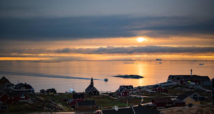 Petite ville côtière groenlandaise au coucher du soleil avec des bateaux traversant les eaux dorées et des icebergs au loin.