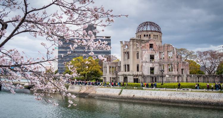 Edificio con cúpula arruinado del Monumento Conmemorativo de la Paz de Hiroshima enmarcado por cerezos en flor a lo largo de un paseo junto al río.