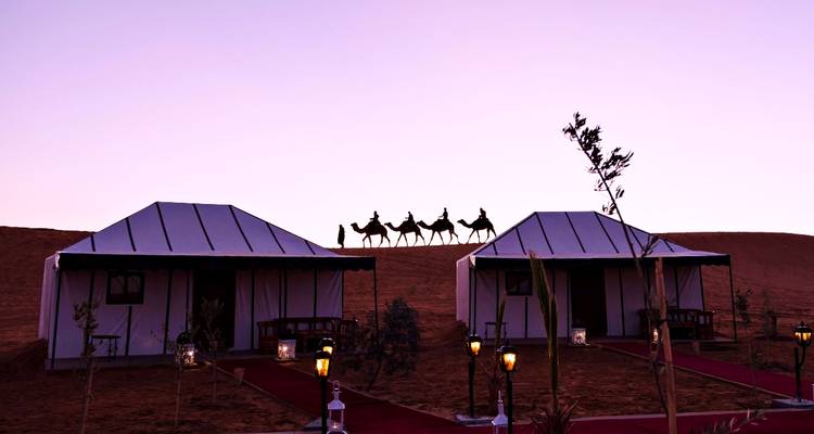Tiendas de lujo en el desierto bajo una cresta donde una fila de camellos se recorta contra un cielo crepuscular violeta