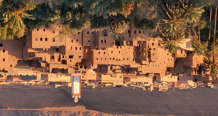 Pueblo de adobe iluminado por el sol con un minarete blanco y alto enmarcado por palmeras y montañas desérticas