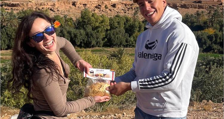 Two travellers laughingly present a bag of snacks with an oasis and cliffs behind them.