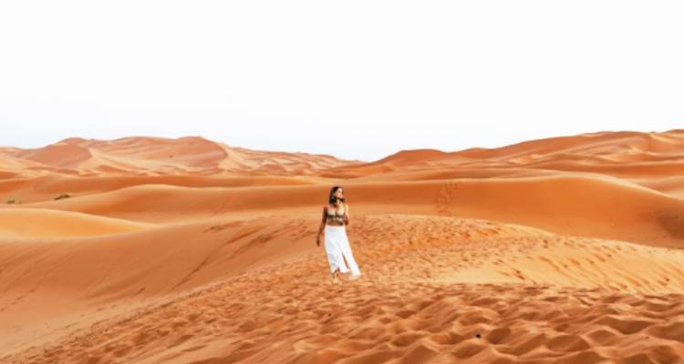 Woman walking alone across rolling golden sand dunes beneath a clear white sky
