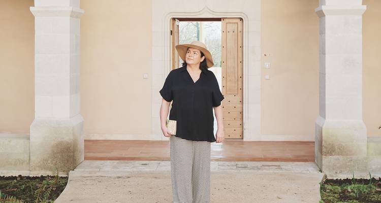 Woman in wide-brimmed hat stands in an elegant chateau courtyard entrance