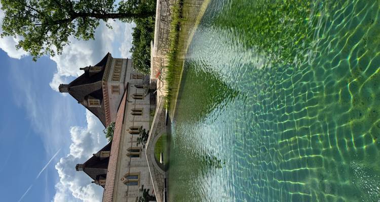 Turquoise natural swimming pond reflects a stone chateau and arching bridge under a blue sky