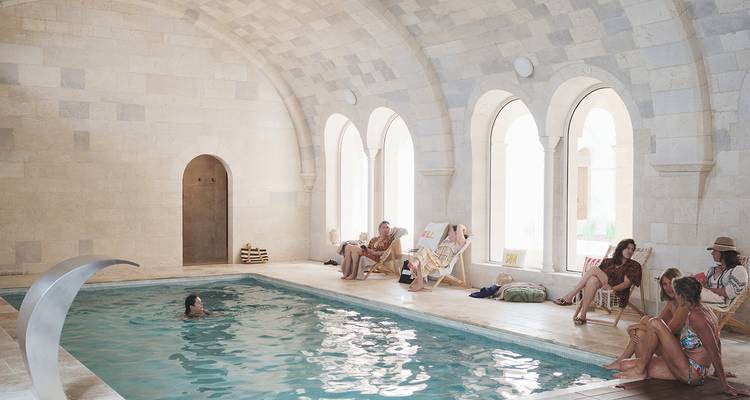 Guests relax on loungers beside an indoor stone pool beneath arched windows