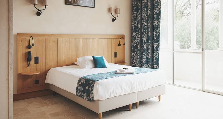 Minimalist hotel room with wooden headboard, large bed, and natural daylight through glass doors.