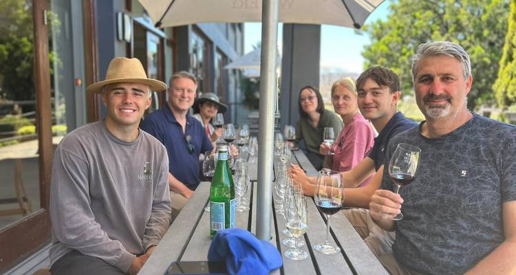 Un petit groupe savourant une dégustation de vin à une longue table en plein air ombragée par des parasols.