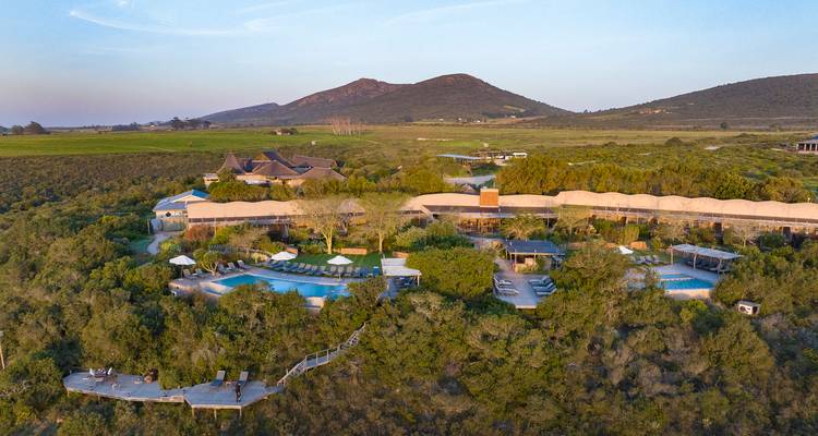Vue aérienne d'un complexe de lodge de luxe avec piscines situé dans le fynbos côtier luxuriant près de collines ondulantes au coucher du soleil.