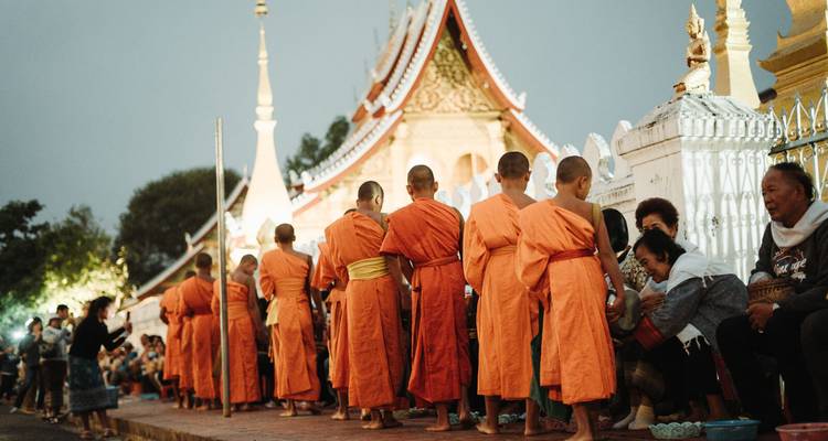 Lange Reihe von orange gekleideten buddhistischen Mönchen, die bei Tagesanbruch neben einem erleuchteten Tempel Almosen erhalten.