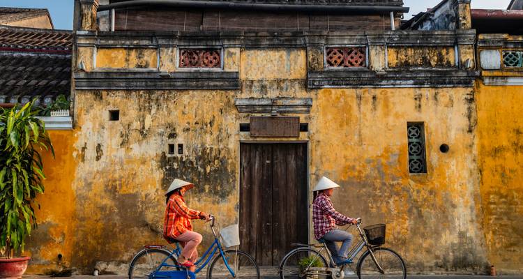 Two cyclists wearing conical hats ride past a weathered yellow wall in Hoi An’s old town.