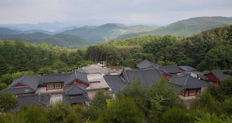 Cluster of grey-tiled temple roofs nestled among pine forested mountains under a hazy sky.