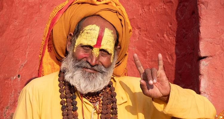 Retrato de un sadhu sonriente con pintura facial amarilla y cuentas de rudraksha posando frente a una pared roja en Nepal.