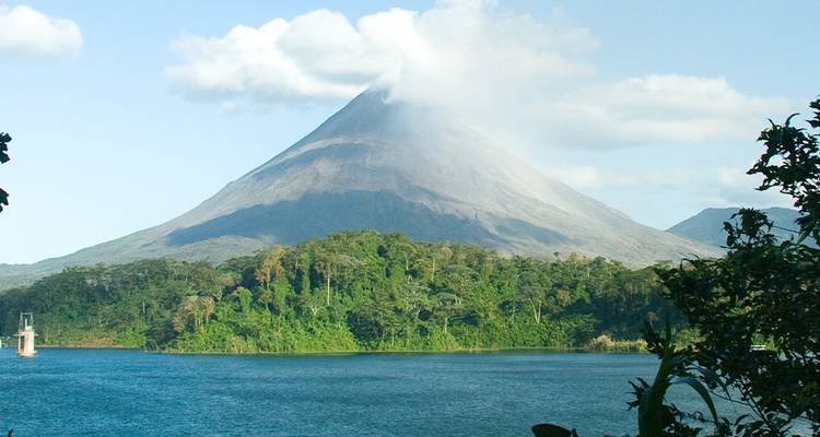 Majestic Arenal Volcano rises above a blue lake, its summit shrouded in a plume of white cloud.