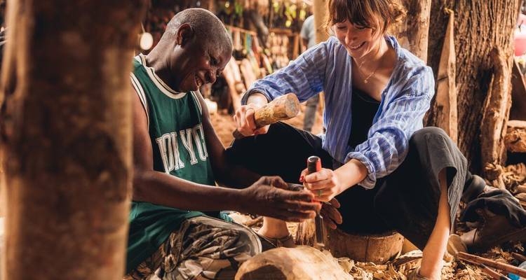 Un voyageur et un artisan local sourient en sculptant le bois ensemble dans un atelier rustique.