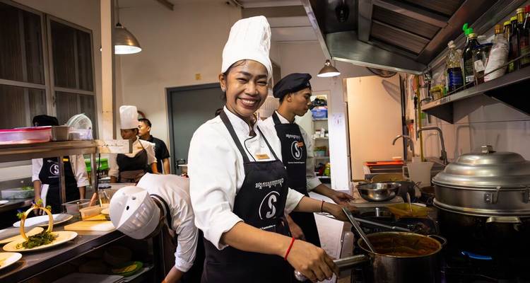 Glimlachende chef-kok in witte hoed leidt een drukke trainingskeuken waar studenten gerechten bereiden.