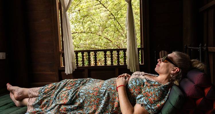 Vrouw in bloemenprint jurk ontspant op een houten daybed naast een open raam omlijst door jungle vegetatie.