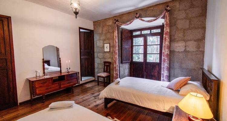 Habitación rústica con dos camas individuales, paredes de piedra, piso de madera y una gran ventana con contraventanas.