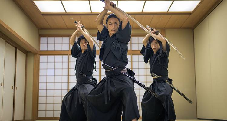 Drie vechtkunstenaars in zwarte hakama demonstreren gesynchroniseerde katanamoves in een tatami dojo.