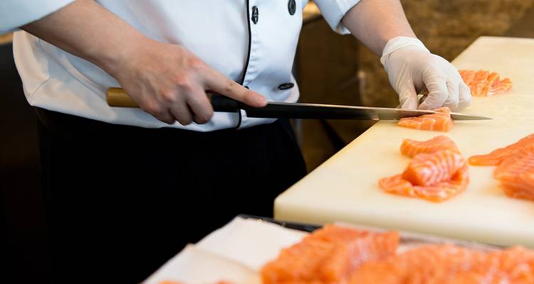 Chef snijdt verse sashimi van zalm met een lang mes op een wit snijplank.