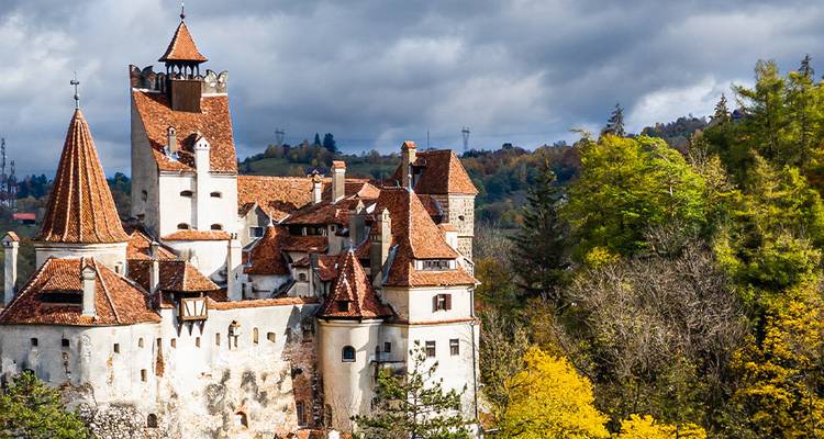 Iconic Bran Castle rises above autumnal forest under dramatic cloudy skies.