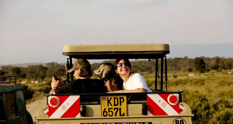 Reizigers glimlachen terwijl ze in een open safari-jeep staan die door grasrijke vlaktes rijdt.