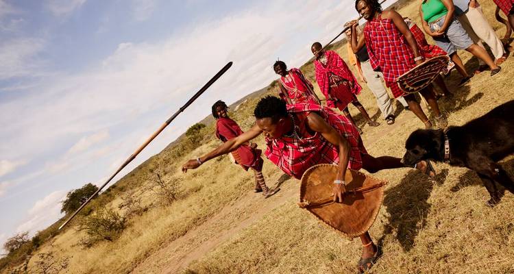 Maasai-krijger in rood geruite stof gooit een speer in een dynamische culturele demonstratie terwijl toeschouwers toekijken.