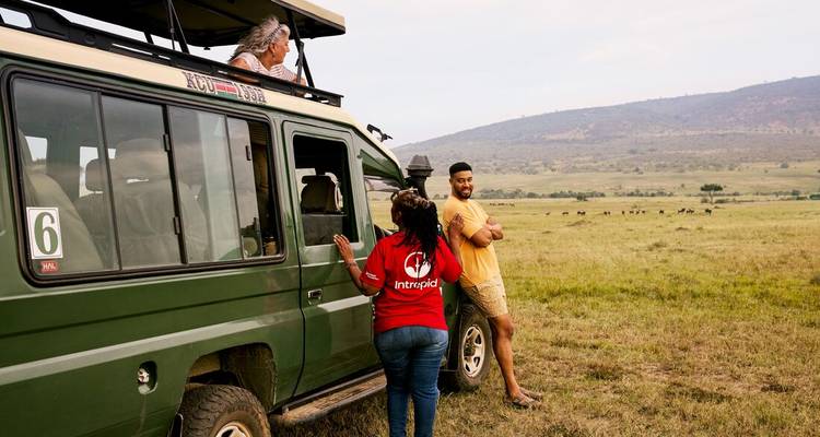 Reizigers chatten naast een groene safariauto met uitzicht op een brede Afrikaanse savanne.