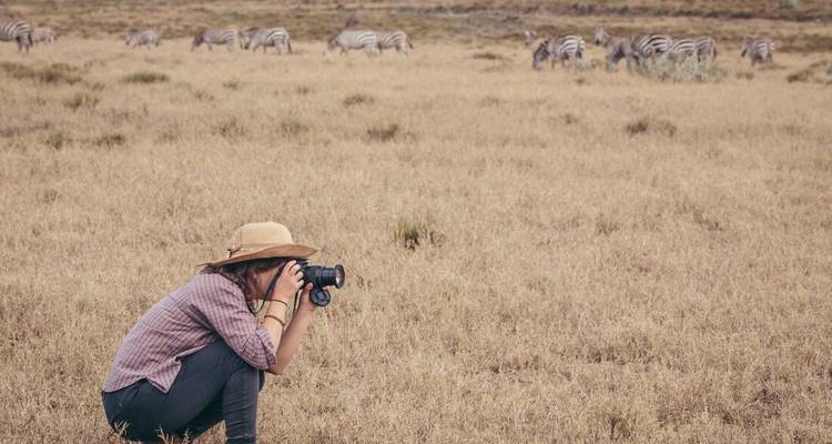 Een jonge reiziger hurkt in droog savannegrasveld en fotografeert een verre kudde zebra's.