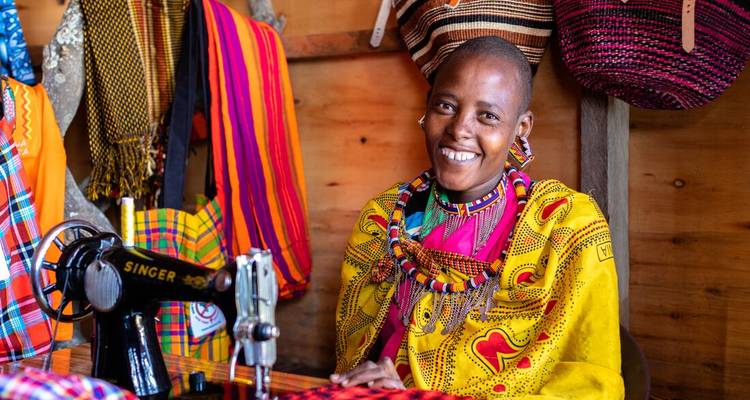 Een glimlachende Maasai-vrouw in kleurige traditionele kleding werkt aan een vintage naaimachine in een ambachtskraam.